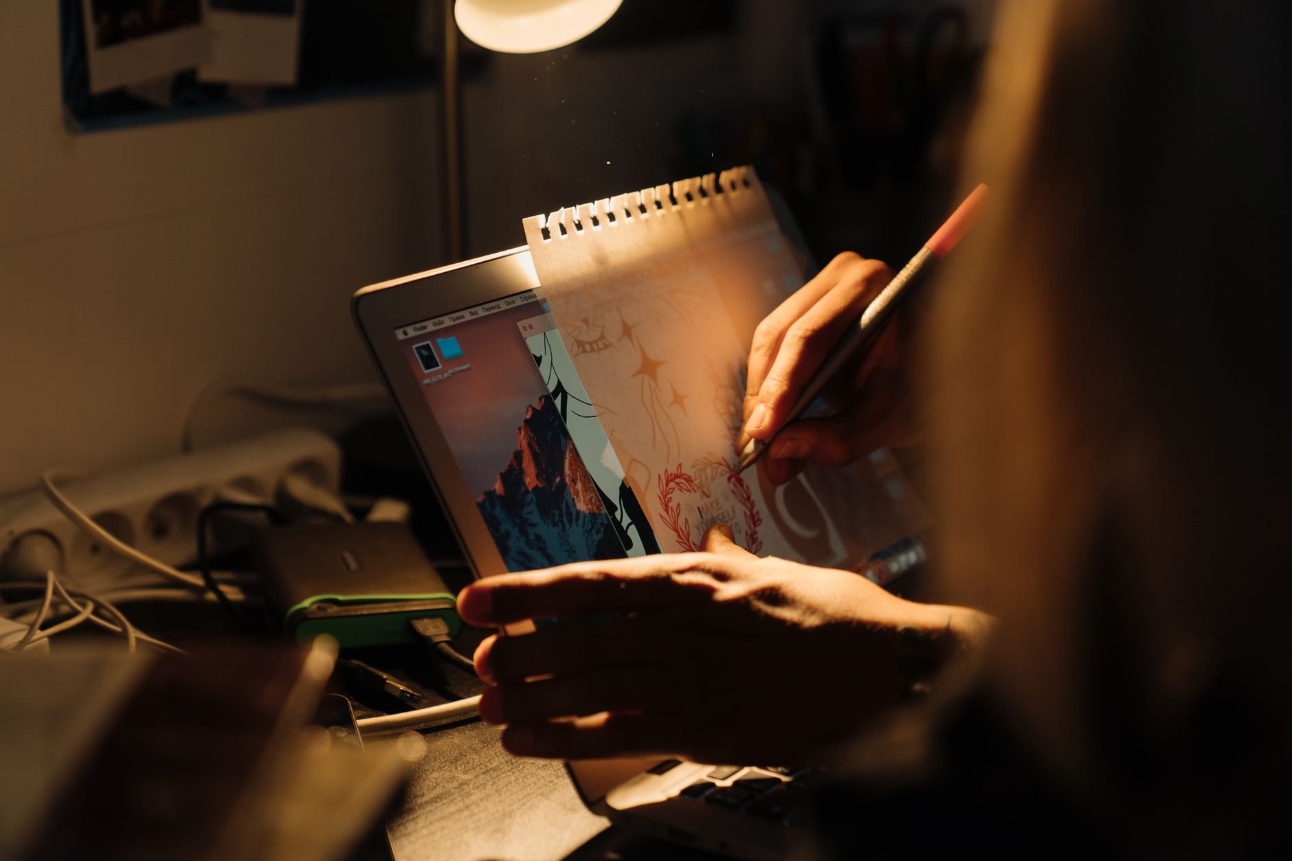 Artist working on a design using a laptop and paper under warm lighting